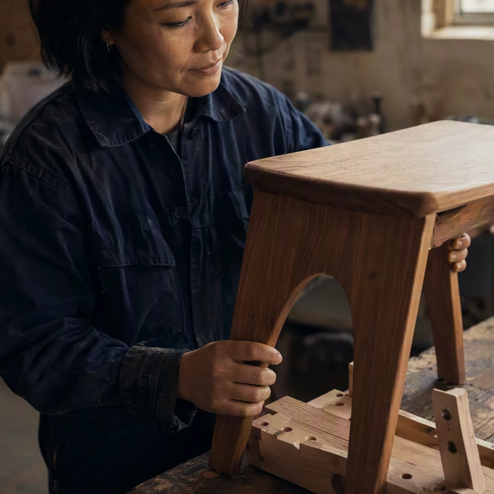 A craftsperson checks a wooden stool beside its assembly jig.